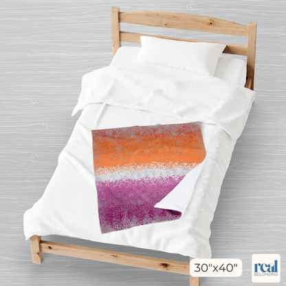 Colorful 30"x40" velveteen plush blanket with lesbian pride color spots dancing across a gray background, laid out on one half of a full-size bed. The bottom corner is folded up to show the white underside of the blanket.