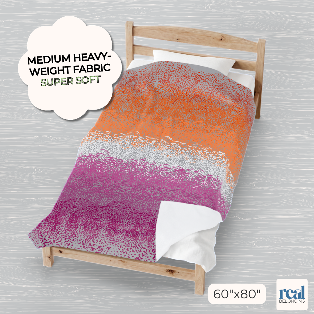 Colorful 60x80" velveteen plush blanket with lesbian pride color spots dancing across a gray background, laid out on a full-size bed with the edges draped comfortably to the sides and one corner folded up to show the solid white underside.